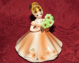 May include: A porcelain figurine of a girl in a peach-colored dress holding a basket of green and yellow flowers.