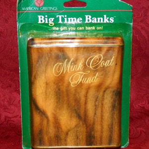 May include: A rectangular tin bank with a faux fur design, labeled "Mink Coat Fund." The bank is packaged in a green plastic blister pack with the text "Big Time Banks" and "American Greetings."