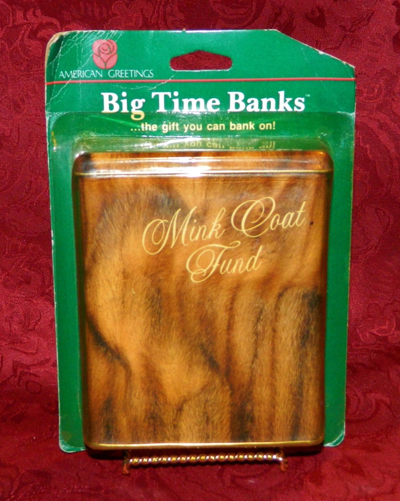 May include: A rectangular tin bank with a faux fur design, labeled "Mink Coat Fund." The bank is packaged in a green plastic blister pack with the text "Big Time Banks" and "American Greetings."