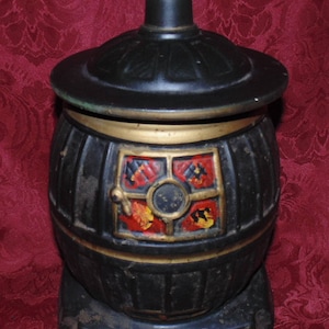Puede incluir: Jarra de galletas de cerámica negra y dorada con forma de estufa de leña con un diseño pintado en rojo, amarillo y negro en la parte delantera.