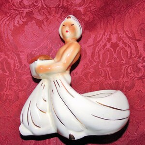 Puede incluir: Un macetero de cer&aacute;mica blanca con forma de mujer que lleva un turbante y sostiene un cuenco. El macetero tiene detalles dorados y est&aacute; decorado con un esmalte marr&oacute;n.