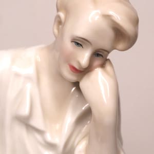 Op de afbeelding: Een wit porseleinen beeldje van een persoon met kort, golvend haar, die een wit shirt draagt, met het hoofd rustend op de hand, naar beneden kijkend.