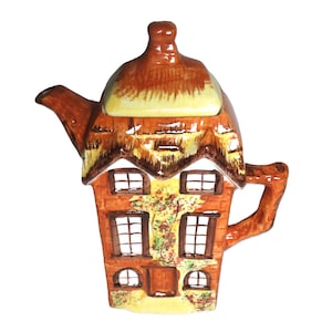 Op de afbeelding: Een keramische theepot in de vorm van een huisje. De theepot heeft een bruin rieten dak, bakstenen muren en meerdere ramen. Het heeft een handvat en een tuit. De theepot is beschilderd met verschillende kleuren.