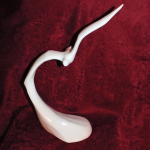 Op de afbeelding: Witte keramische sculptuur van een vogel in vlucht. De vogel bevindt zich in een sierlijke, gebogen houding met uitgespreide vleugels. De sculptuur staat op een rode achtergrond.