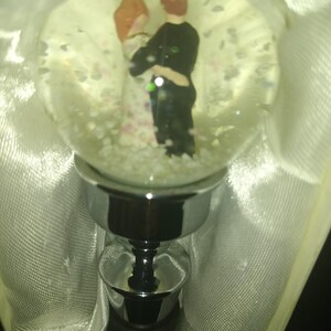May include: A decorative bottle stopper featuring a clear glass globe with a couple figurine inside. The couple is embracing, surrounded by small, white confetti-like shapes. The stopper has a silver-colored metal base and a black rubber seal.