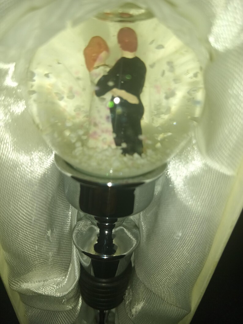 May include: A decorative bottle stopper featuring a clear glass globe with a couple figurine inside. The couple is embracing, surrounded by small, white confetti-like shapes. The stopper has a silver-colored metal base and a black rubber seal.