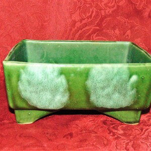 Puede incluir: Un macetero de cerámica verde con forma cuadrada y cuatro patas. El macetero tiene un esmalte verde moteado con acentos blancos.