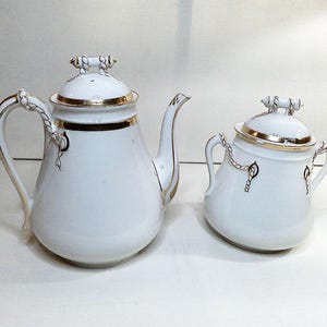 May include: A white porcelain tea set with gold trim. The set includes a teapot and a sugar bowl, both with rope-like handles and lids. The teapot has a curved spout. The set is likely vintage.
