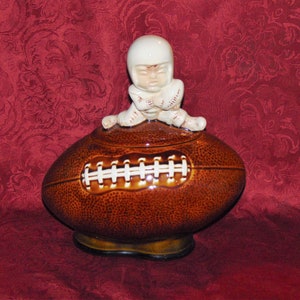 Puede incluir: Un tarro de galletas de cerámica marrón y blanco en forma de balón de fútbol con una pequeña figura de jugador de fútbol en la parte superior. El jugador lleva un casco y una camiseta blancos.