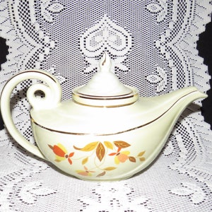 Op de afbeelding: Een vintage theepot in de vorm van een djinn-lamp met een crèmekleurig lichaam en gouden afwerking. De theepot heeft een gestileerde handgreep en tuit en heeft een bloemenpatroon met oranje, gele en bruine bladeren.