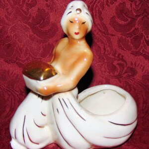 Puede incluir: Una figurita de cer&aacute;mica blanca de una mujer con un turbante blanco con un acento dorado. Ella sostiene un taz&oacute;n dorado en sus manos. La figurita es una maceta con una maceta de cer&aacute;mica blanca.