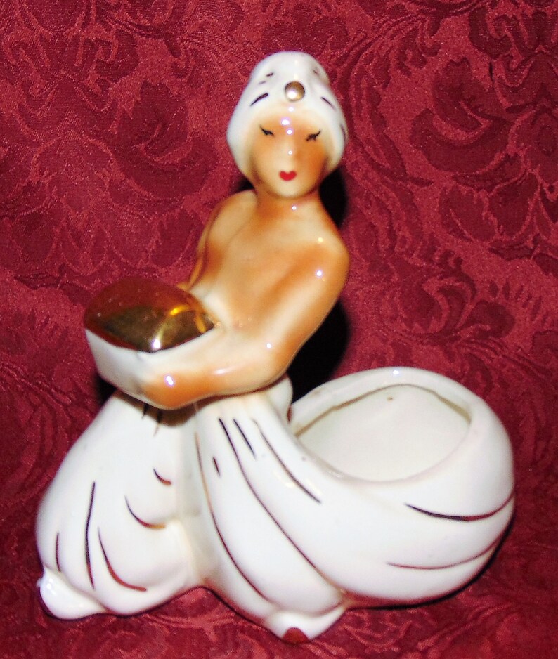 Puede incluir: Una figurita de cer&aacute;mica blanca de una mujer con un turbante blanco con un acento dorado. Ella sostiene un taz&oacute;n dorado en sus manos. La figurita es una maceta con una maceta de cer&aacute;mica blanca.