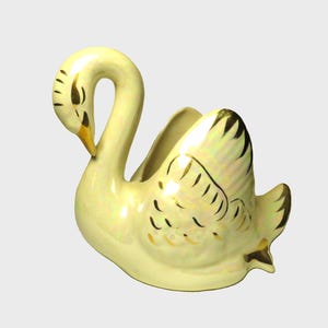 May include: A white ceramic swan planter with gold accents. The swan has a detailed design on its wings and a gold beak.