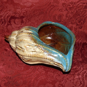 May include: A ceramic seashell planter with a blue and brown glaze. The planter has a textured, white exterior and a smooth, glazed interior.