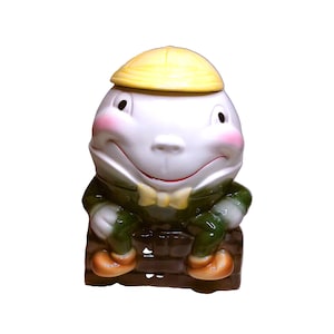 Könnte beinhalten: Eine Keramikfigur von Humpty Dumpty mit gelbem Hut, rosigen Wangen und grüner Jacke. Die Figur sitzt auf einer braunen Backsteinmauer und hat orangefarbene Schuhe. Das Design ist skurril und dekorativ.