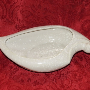 May include: A white ceramic planter shaped like a bird with a speckled glaze. The planter has a wide, shallow bowl and a curved spout.