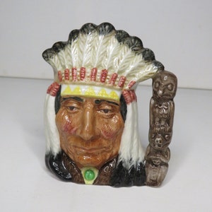 Pode incluir: Uma figurinha de cerâmica de um chefe nativo americano usando um cocar de penas. A figurinha tem um rosto marrom com uma pedra verde ao redor do pescoço. A figurinha está segurando um totem com entalhes de animais.