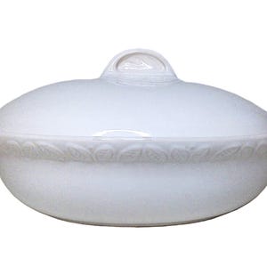 Könnte beinhalten: Weiße ovale Servierschüssel mit Deckel und Griff. Die Schüssel hat ein dekoratives Blättermuster am Rand. Der Deckel hat einen kleinen, gewölbten Griff. Die Schüssel ist aus Keramik.