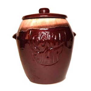 Könnte beinhalten: Ein Vintage-Keramiktopf mit Deckel. Der Topf hat eine tief burgunderrote Farbe mit einem helleren braunen und beigen Band um den oberen Rand. Der Topf hat ein erhabenes Design auf der Vorderseite und zwei kleine Griffe an den Seiten.