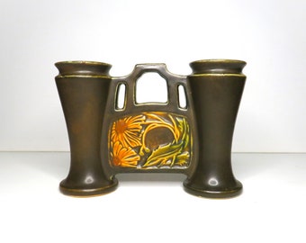 May include: A brown ceramic double vase with a floral design in shades of yellow, green, and brown. The vase has two openings for flowers and a decorative cutout in the center.