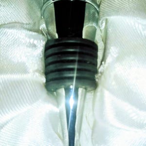 May include: A close-up of a silver wine bottle stopper with a black rubber seal. The stopper has a decorative, ridged design and is set against a white, textured background.