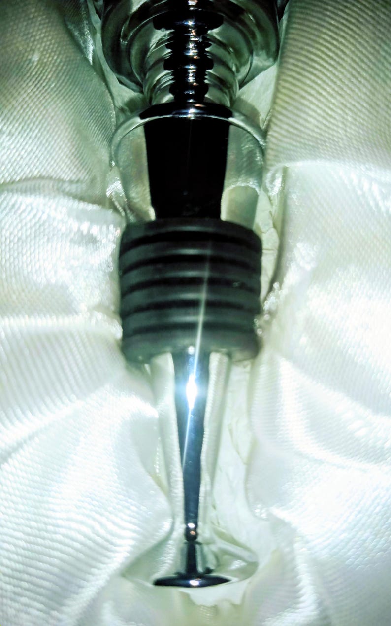 May include: A close-up of a silver wine bottle stopper with a black rubber seal. The stopper has a decorative, ridged design and is set against a white, textured background.