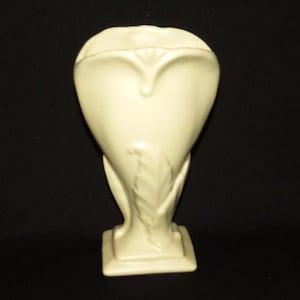 Könnte beinhalten: Eine cremefarbene Keramikvase mit einem stilisierten Blumendesign. Die Vase hat eine herzförmige Öffnung und einen quadratischen Sockel.