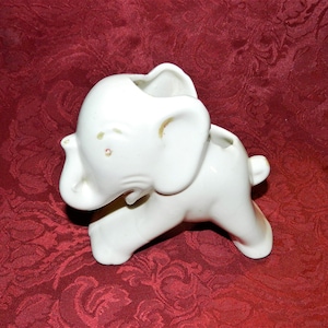 May include: White ceramic elephant planter with a small opening on the top for planting. The elephant is standing on all four legs with its trunk raised.