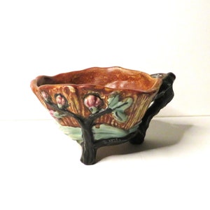 May include: A brown and orange ceramic bowl with a tree branch handle and three legs. The bowl features a textured design with pink flowers and green leaves. The handle and legs are dark brown.