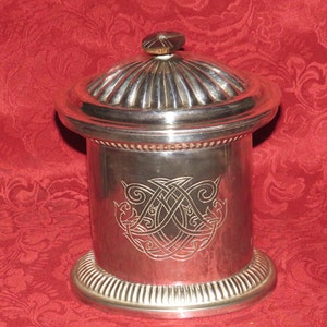Puede incluir: Un recipiente cilíndrico de plata con una tapa abovedada y un intrincado diseño de nudos celtas. El recipiente tiene un borde con cuentas y una tapa acanalada.