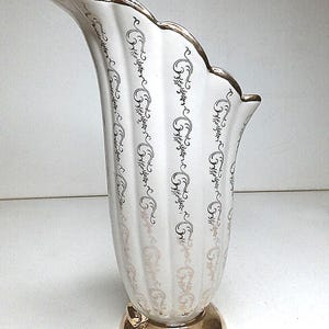 Könnte beinhalten: Eine weiße Keramikvase mit goldfarbenem Rand und aufwendigen goldenen Verzierungen. Die Vase hat eine einzigartige, geschwungene Form und eine breite Öffnung.