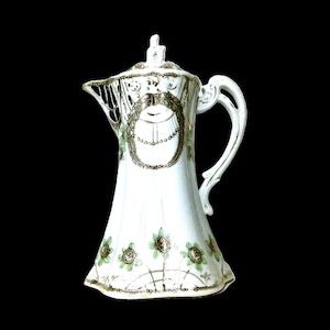 May include: White porcelain coffee pot with a decorative design. The pot has a detailed handle, a lid with a finial, and a spout with an openwork design. The body is decorated with gold and green floral accents and a central oval design.
