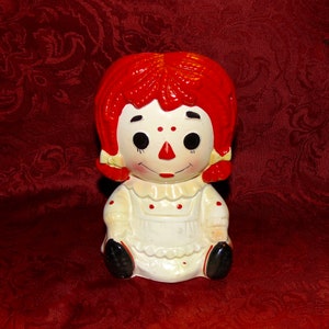 May include: A vintage ceramic Raggedy Ann doll piggy bank with red hair, a white dress with red polka dots, and black shoes.