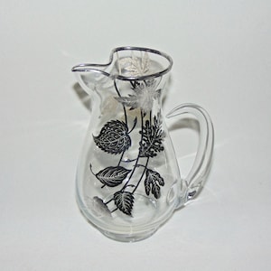 Puede incluir: Una jarra de vidrio transparente con un diseño de hojas plateadas. Las hojas son negras y plateadas y están dispuestas en forma de vid.