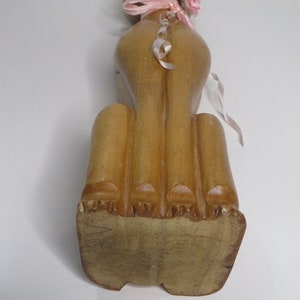 Puede incluir: Una figurita de gato de madera con una cinta rosa y una flor alrededor del cuello. El gato est&aacute; sentado derecho con la cola enroscada alrededor de su cuerpo. La figurita est&aacute; hecha de madera marr&oacute;n claro y tiene un acabado liso.
