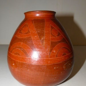 May include: A red clay pot with a geometric design in a darker shade of red. The pot has a rounded body and a narrow opening.