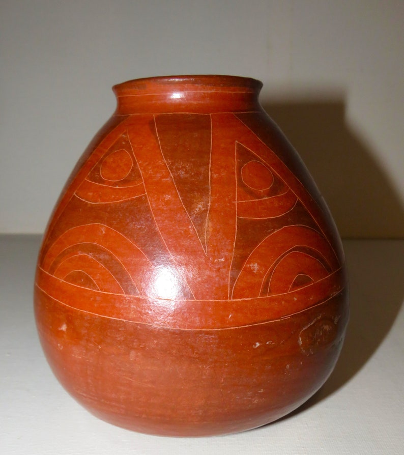 May include: A red clay pot with a geometric design in a darker shade of red. The pot has a rounded body and a narrow opening.