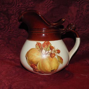 May include: A brown and white ceramic jug with a handle and a pear design. The jug has a brown glaze on the top and a white glaze on the bottom. The pear design is in shades of yellow, orange, and green.