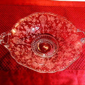May include: A clear glass serving dish with a floral pattern and two handles. The dish has a scalloped edge and a circular indentation in the center. The dish is on a red surface.