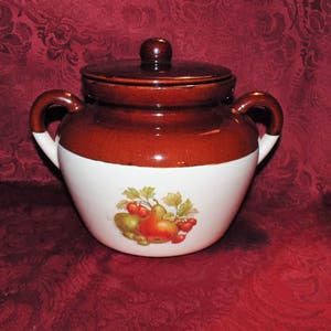 Puede incluir: Una olla de cerámica marrón y blanca con tapa, que presenta un diseño de frutas con peras, manzanas y fresas. La olla tiene dos asas y está decorada con una banda blanca alrededor del centro.