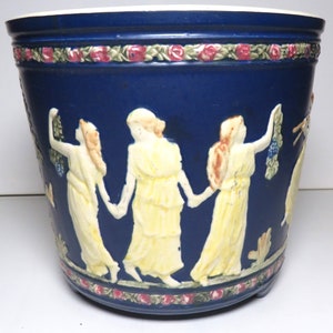 May include: A blue ceramic planter with a raised design of three women in white dresses holding garlands. The planter has a floral border around the top and bottom.
