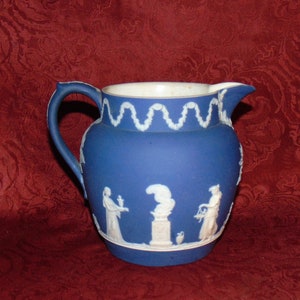May include: A blue and white ceramic pitcher with a white design of figures and a garland around the top.