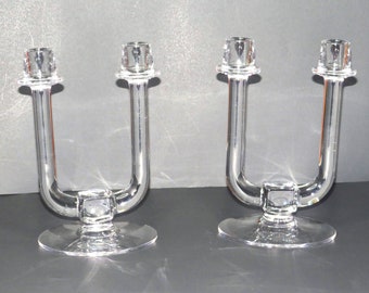May include: A pair of clear glass candle holders with a U-shaped design. The holders have a round base and a square-shaped centre.