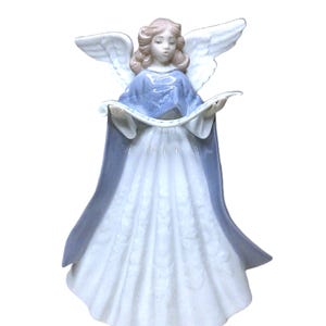 May include: A ceramic angel figurine, featuring a blue and white gown and holding a scroll. The angel has white wings and pink hair. A decorative item for home decor, suitable for display.