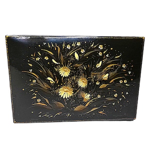 Puede incluir: Una caja rectangular negra con un diseño floral dorado. Las flores son estilizadas y tienen un aspecto delicado e intrincado. La caja está hecha de madera y tiene un acabado brillante.