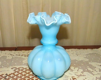 May include: A light blue glass vase with a ruffled top and a round, bulbous body. The vase is sitting on a white crocheted doily.