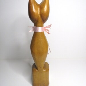 Puede incluir: Una figurita de gato de madera con una cinta rosa alrededor del cuello. El gato est&aacute; de pie sobre una base de madera.