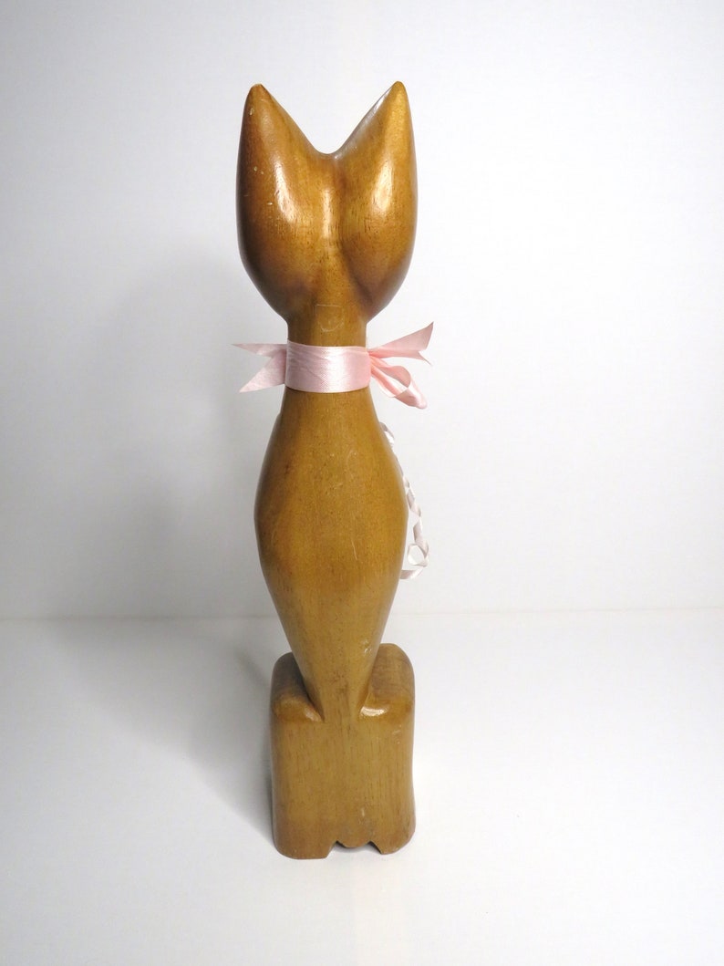 Puede incluir: Una figurita de gato de madera con una cinta rosa alrededor del cuello. El gato est&aacute; de pie sobre una base de madera.