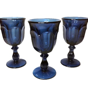 May include: Three cobalt blue stemmed water glasses. Each glass has a tulip-shaped bowl and a decorative stem. The glasses are made of a dark blue glass and are displayed against a white background.