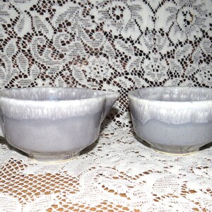 May include: Two ceramic bowls in a light purple glaze with a white rim. The bowls are on a white lace tablecloth.